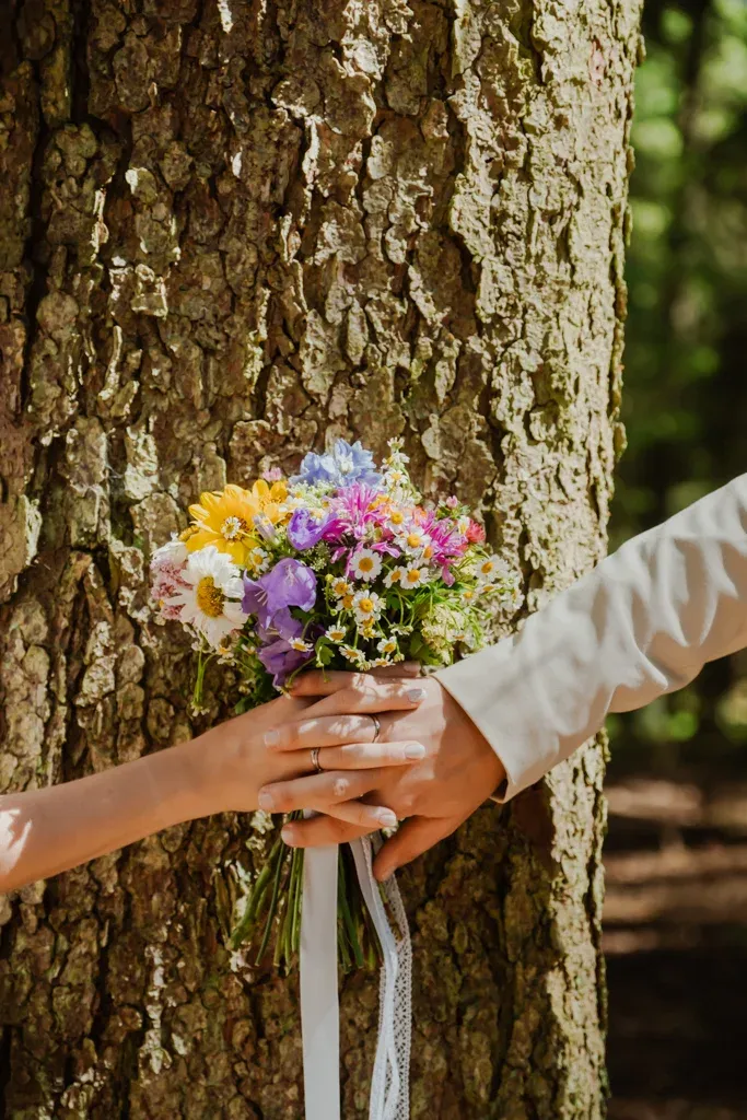 Hände eines Brautpaars mit Eheringen halten einen Wiesenblumenstrauß vor einem Baumstamm
